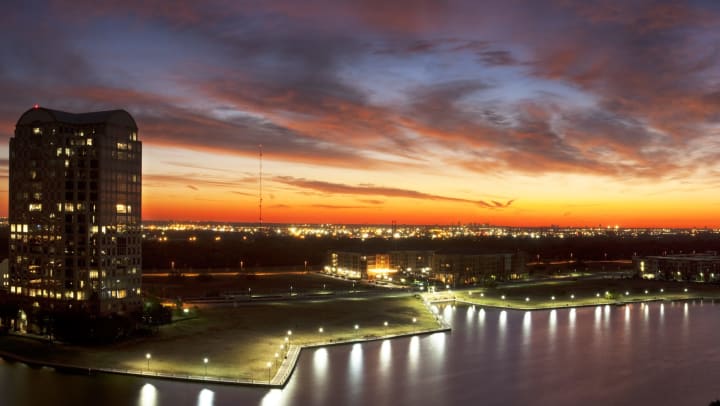 A landscape view of water, green space, buildings, and city lights in the distance, with a colorful sunset in the sky.