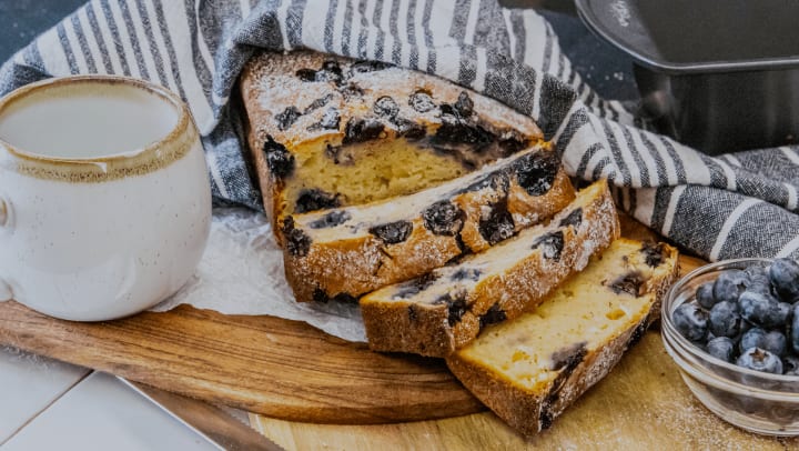 Blueberry loaf displayed under a stripe blue, white linen cloth on a wooden board next to a artisan crafted glass mug and a small bowl of blueberries