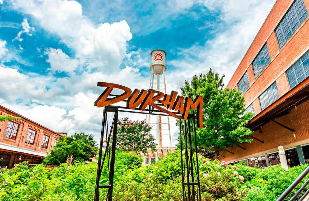 A large, rustic durham sign stands in a lush green garden area near Stride in Durham, North Carolina