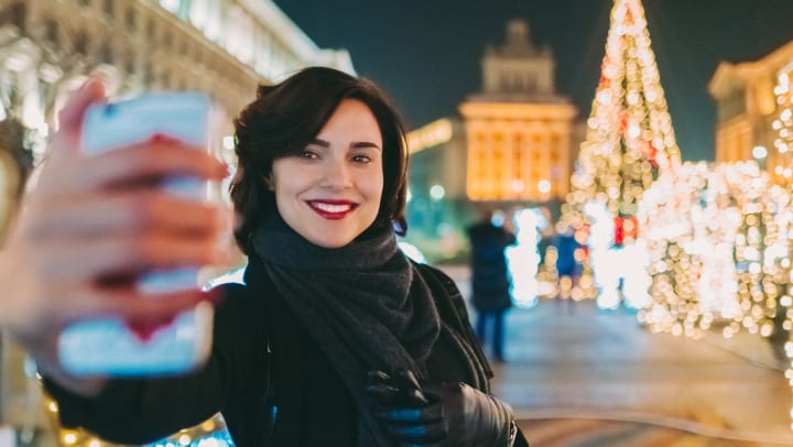 Woman taking a selfie in front of an out-of-focus holiday lights display. 