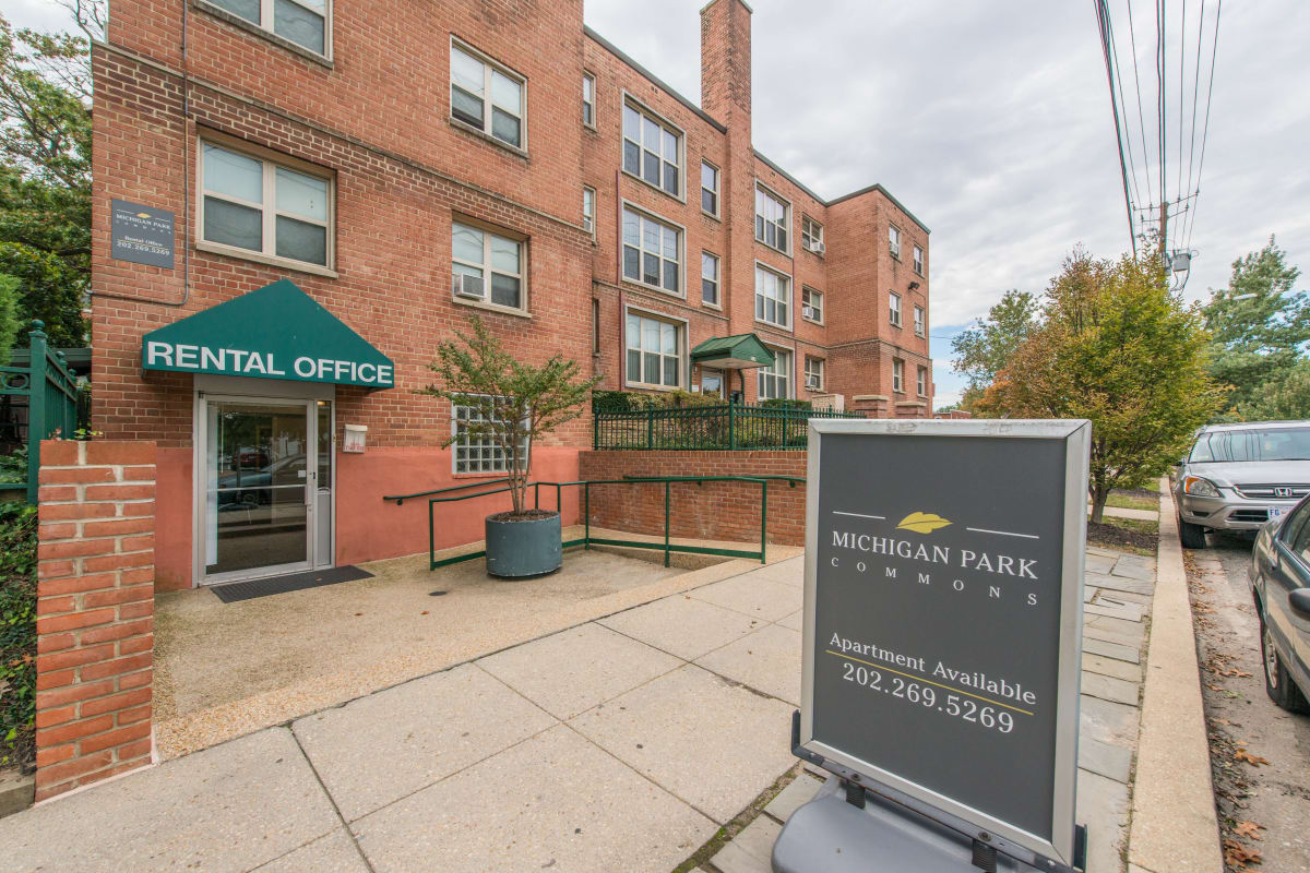 Street view of the modern apartment building with a welcoming sign in front at Michigan Park Commons in Washington, District of Columbia