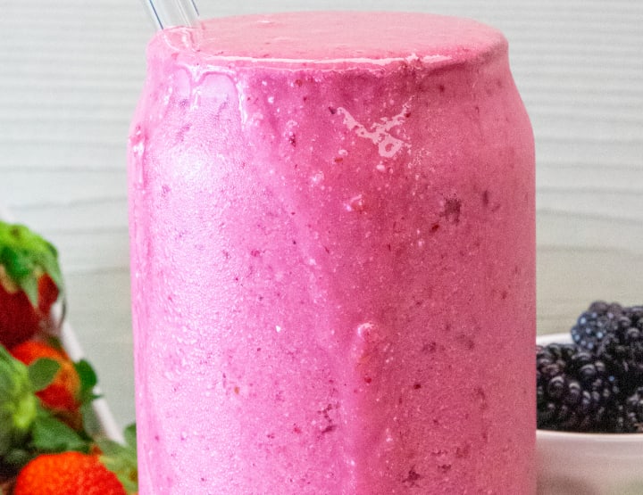 A darkish-pink smoothie in a beer can glass cup with a glass straw inside with a small bowl of blackberries and a bunch of strawberries behind