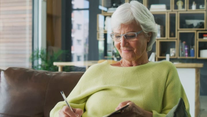Older woman smiling and writing in a notebook while sitting on a couch