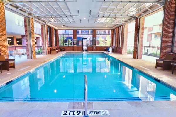 Indoor pool at Watermere Frisco with clear blue water, brick walls, lounge seating, and roll-up garage-style windows.
