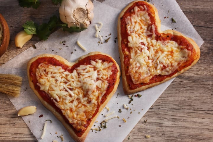 Heart-shaped cheese pizzas presented on parchment paper and a wooden counter-top, surrounded by garlic and fresh basil leaves