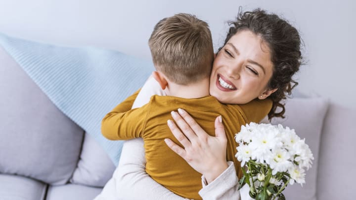  Young boy hugging an adult woman who is smiling and holding a bouquet of white daisies. 