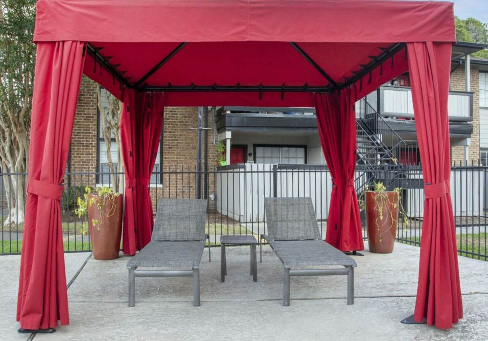 A poolside cabana with red canopy and curtains and two lounge chairs underneath at Rayfords Edge Apartments in Spring, Texas