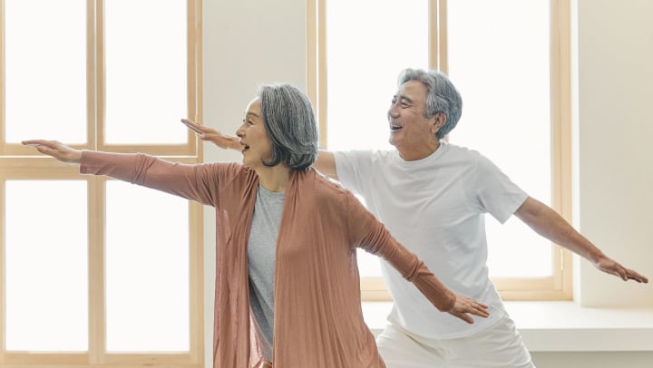 An older couple smiling while stretching during exercise in front of a well-lit window
