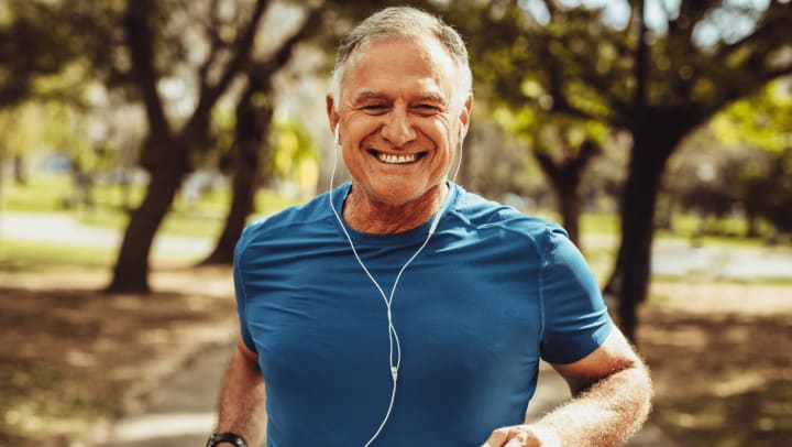 An elderly man running through a park, surrounded by trees and grass, showcasing an active lifestyle.