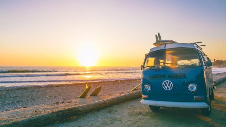A Volkswagen van parked on the beach at sunset with surfboards attached to the roof and along the side of the van.