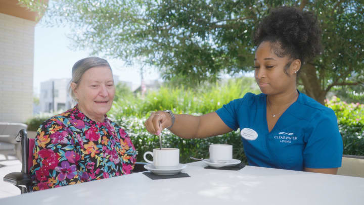A senior Clearwater resident in a floral dress sitting at a table on an outdoor patio having coffee with her care partner wearing her blue uniform.