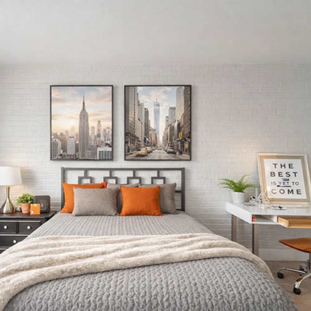 Cozy bedroom with gray bedding, orange accent pillows, and modern artwork on white brick wall at Flats on Fifth in Louisville, Kentucky