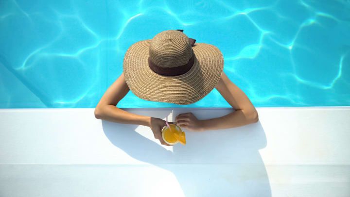 Young woman in elegant hat drinking a cocktail at the edge of a swimming pool