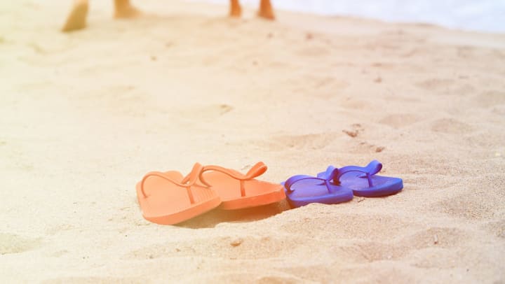 One pair of red sandals and one pair of blue sandals on a sandy beach with two pairs of legs in the background