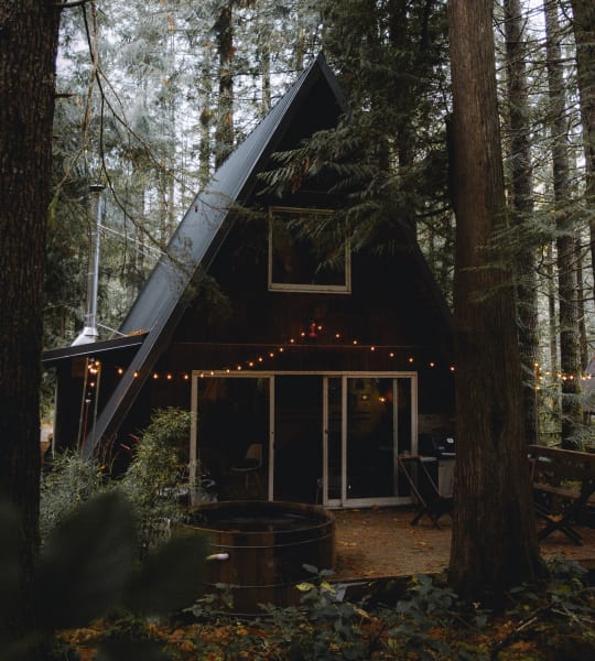 a black a Frame cabin in the woods with a small deck on the front and strung lights around the deck 