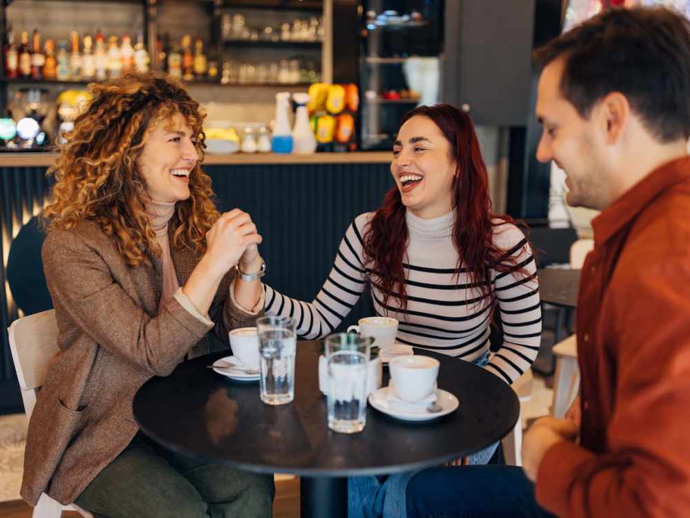 Three friends laughing and enjoying coffee at a stylish cafe table with drinks and pastries at 43 North Apartments in Grand Haven, Michigan