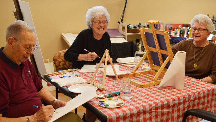 Image of three Touchmark residents smiling as they paint on easels together.
