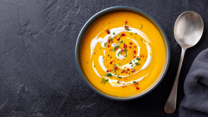 A bowl of pumpkin soup with a white swirl sitting next to a spoon on a dark background. 