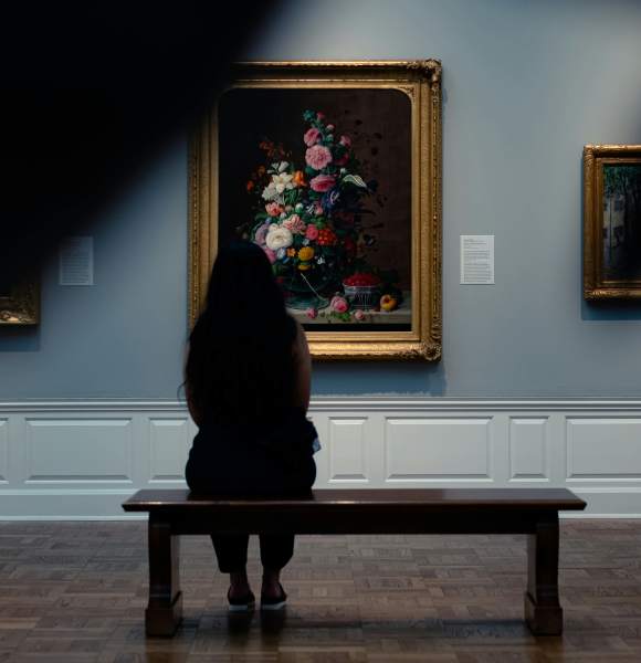 a from behind a person sitting on a bench in an art museum looking at a piece of art in an ornate gold frame