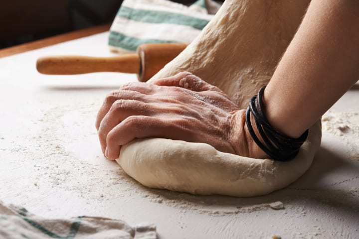 A persons hands kneading raw dough on a table with a rolling pin and flour in the background