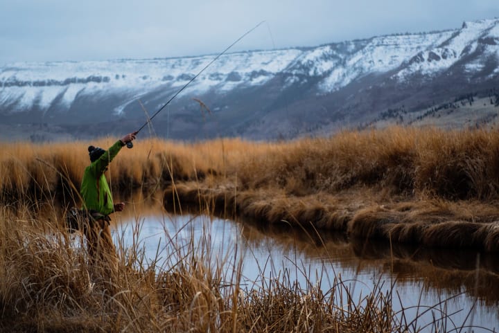 a person stands on the bank of a small river fishing with winter clothing on and snow capped mountains in the background