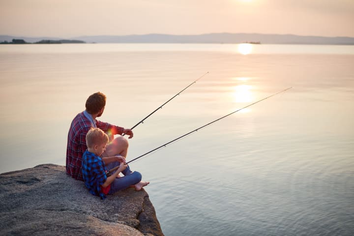 Back view portrait of father and son sitting together on rocks fishing with rods in calm lake waters with landscape of setting sun