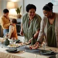 Two women setting a table for dinner while two children hug in the background at Cypress McKinney Falls in Austin, Texas