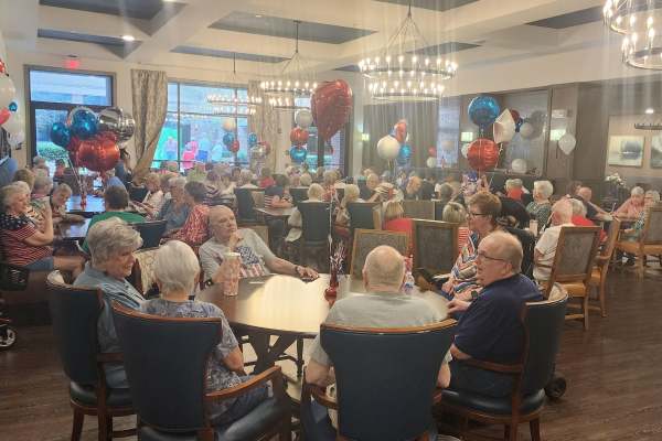 Lively community dining room filled with residents enjoying dinner together at warmly lit tables.