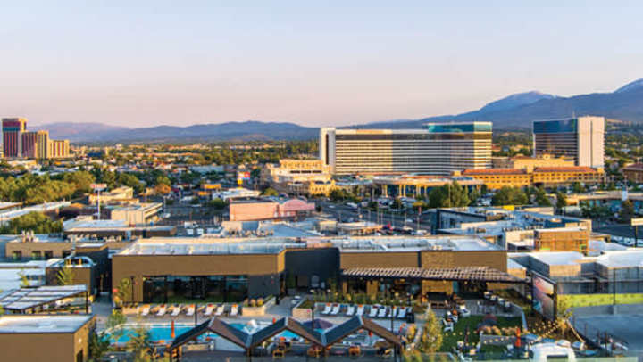 Aerial view of downtown Reno with modern buildings, pools, and outdoor spaces in the foreground, and mountains in the background at sunset.