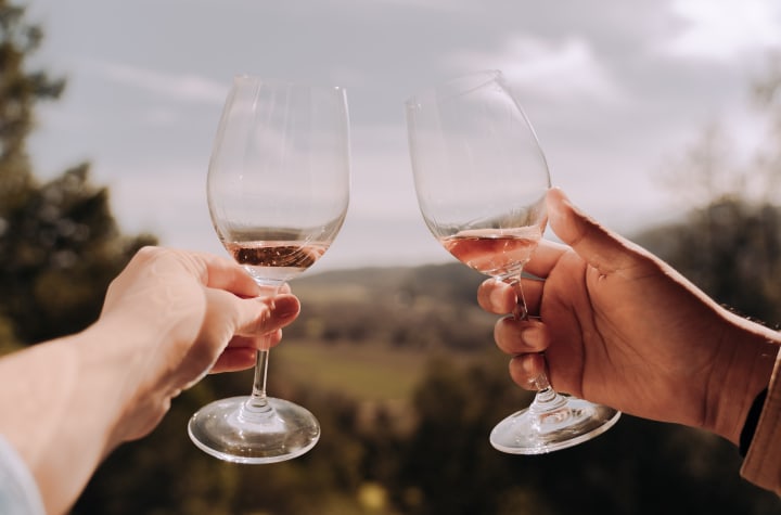 Two hands holding individual wine glasses, half filled with prosecco, overlooking a field of grapes