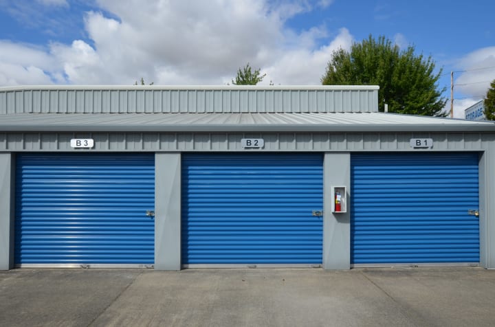 A row of drive up access storage facility doors with a gray building and blue doors on a bright sunny day with blue skies and clouds