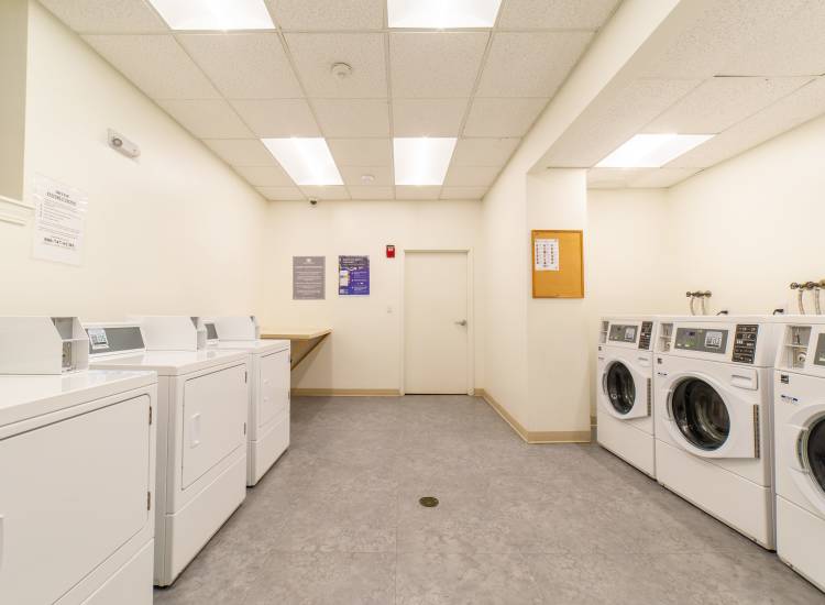Laundry room with white machines, folding table, and informational notices on walls at 15-25 Hemenway Apartments in Boston, Massachusetts