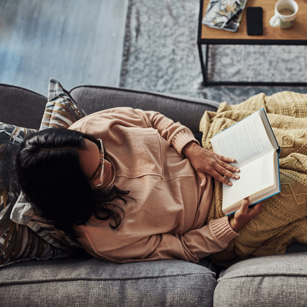 Resident enjoying a book while relaxing on the couch in her apartment home at our Rafael Gardens community at Mission Rock at San Rafael in San Rafael, California