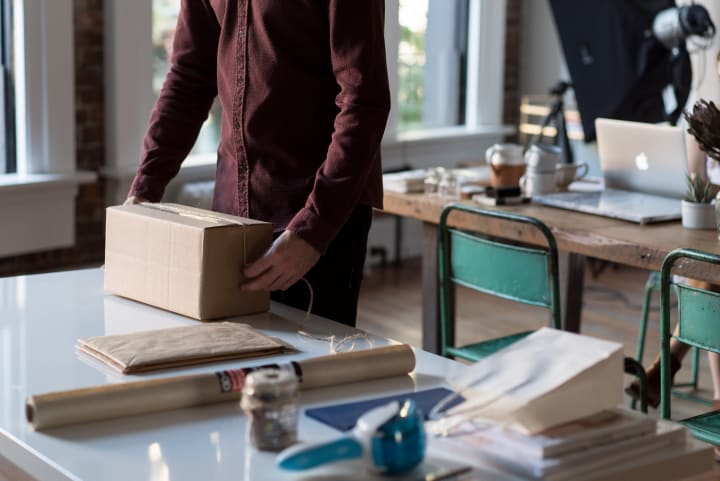 A person from the chest down is packing a box with tape, paper and bags on a table. 