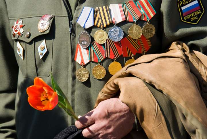 A decorated military veteran wearing a uniform adorned with medals and honors holds a red tulip and a coat, symbolizing service, sacrifice, and remembrance.