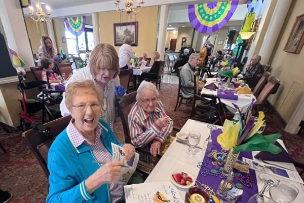 Festive elderly residents gathered at tables in dining room decked out in Mardi Gras decor
