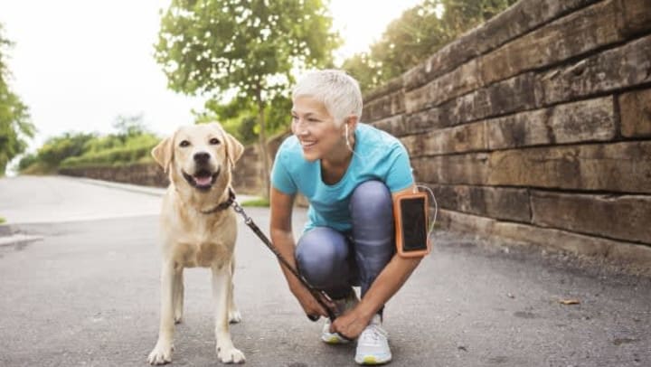 Older woman with headphones on bending at the knee and smiling at her dog