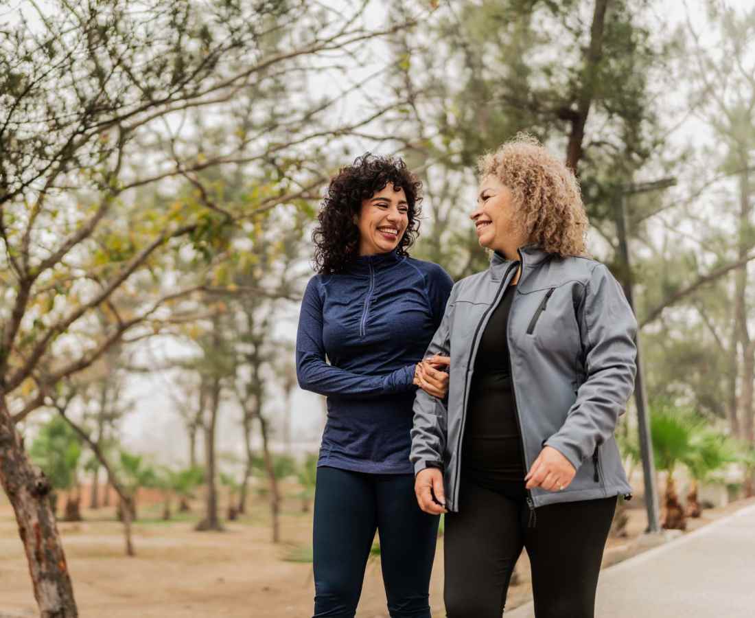 Two women walking together, smiling in a park surrounded by trees and greenery at Valley Brook Townhomes in Rochester, New York