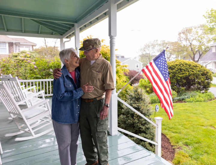 A happy elderly veteran and his spouse embrace on their front porch, smiling with gratitude for their Veterans Discount, as an American flag waves in the background.