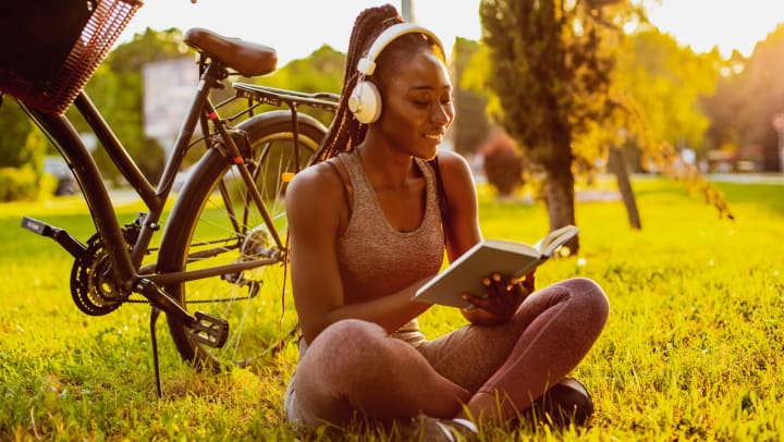 Young woman in athletic clothes wearing headphones and reading a book while sitting on the grass in a park next to a bicycle