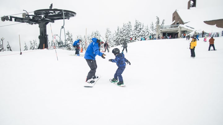 an adult teaching a child to snowboard on a very snowy mountain with other snowboarders and skiers in the background