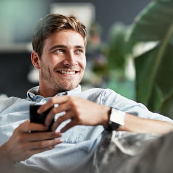 A man sitting on his living room couch with his phone at Cypress Creek at Stoney Ridge in Del Valle, Texas
