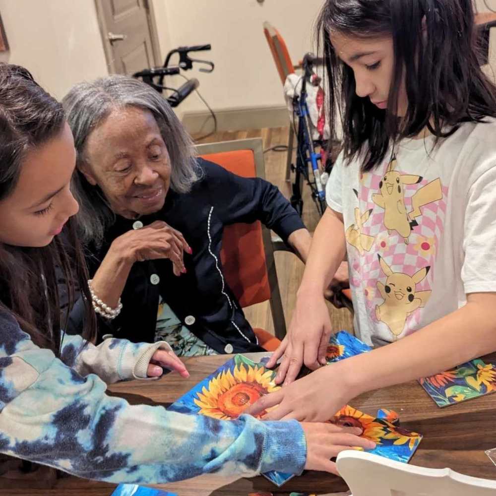 Resident working on a sunflower puzzle with two children during an intergenerational activity at The Landing at Shadow Creek Ranch in Pearland, Texas.