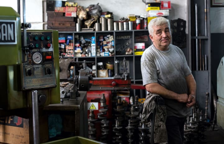 And older man stands in his mechanics shop with his arms crossed and his hands greasy in front of a shelf full of parts and inventory. 