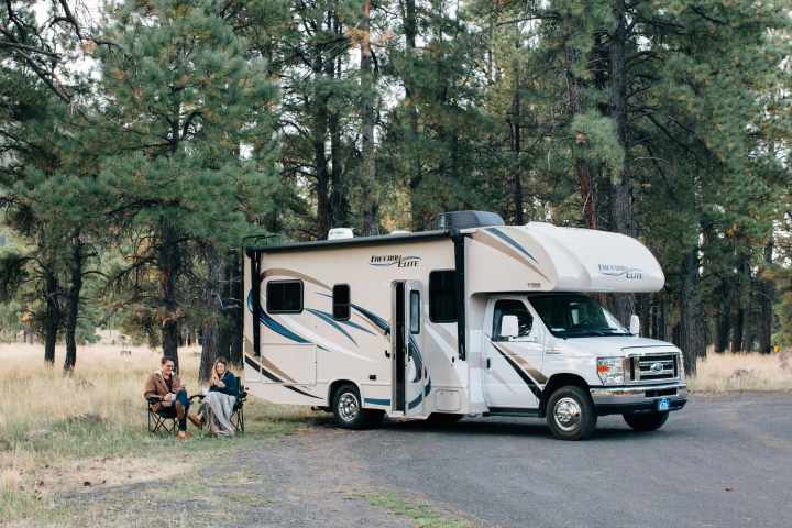 a couple sits in camping chairs with blankets outside of an RV that is parked in the forest 