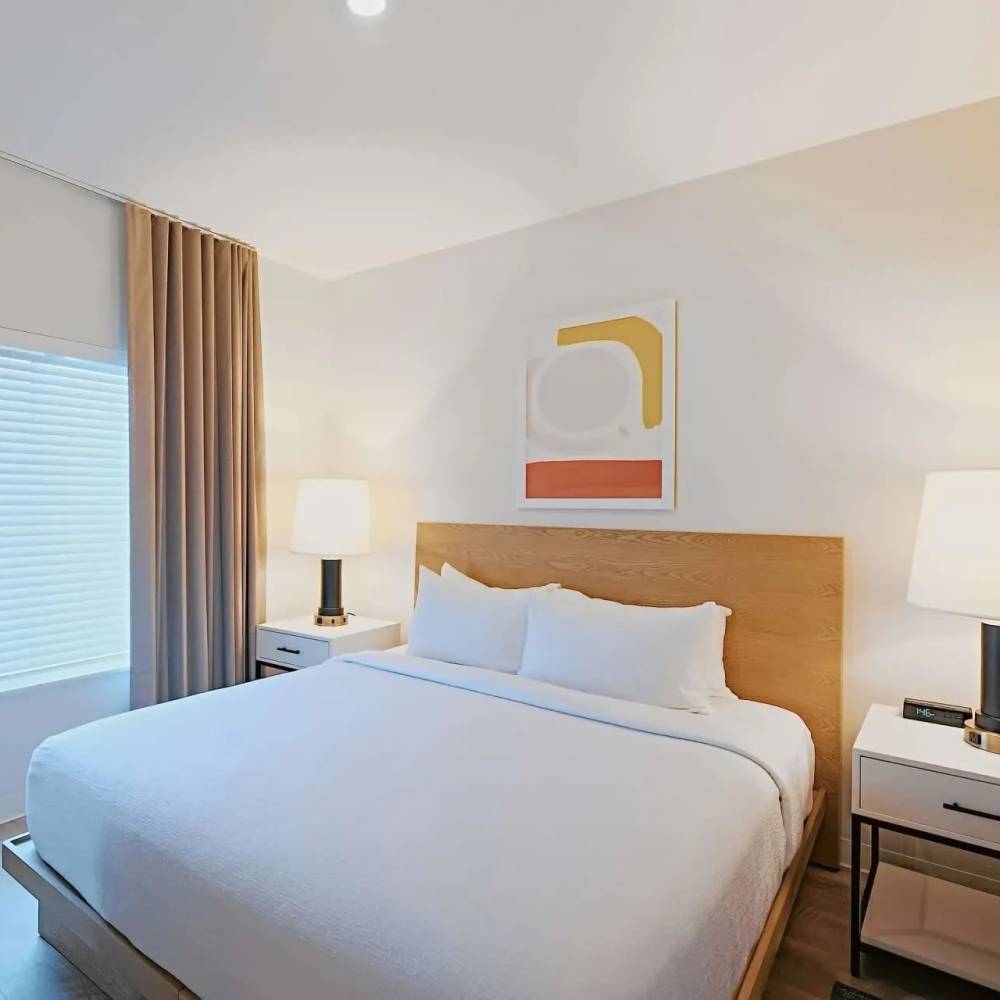 Bright bedroom with a queen bed, modern lamps, and large window with blinds at WaterWalk Boise - Meridian in Meridian, Idaho