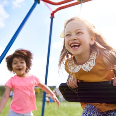  Girl swinging in swing and laughing while another laughing girl watches at playground at Stuart Mesa in Oceanside, California