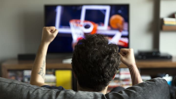 A man on a couch cheering. Basketball playing on the TV in front of him. 