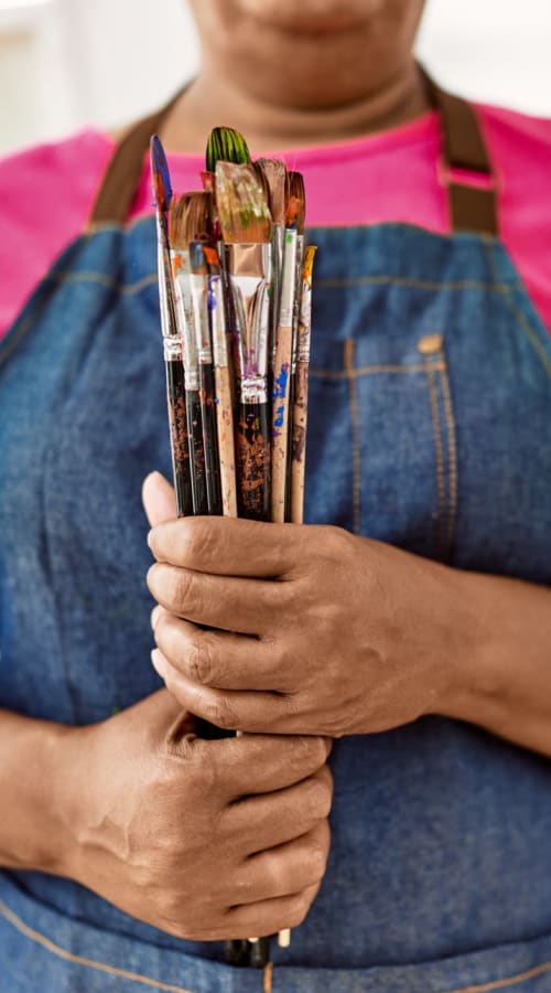 Close up of person wearing an art smock and holding  paint brushes at Pillars Senior Living in Lakeville, Minnesota