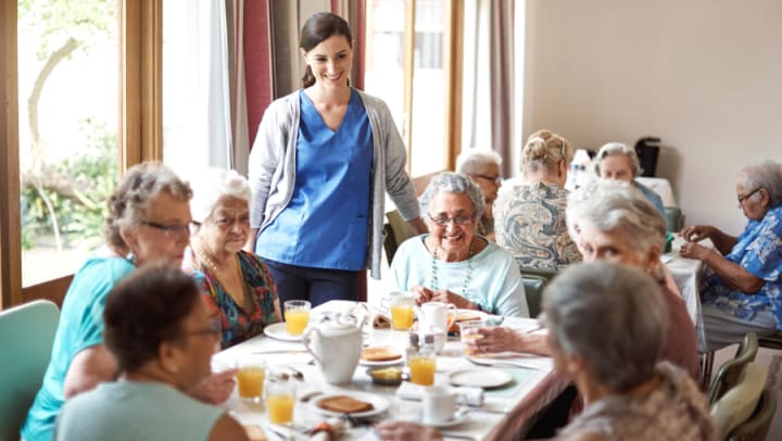 Group of senior ladies having breakfast in a light-filled dining room with a medical assistant nearby.